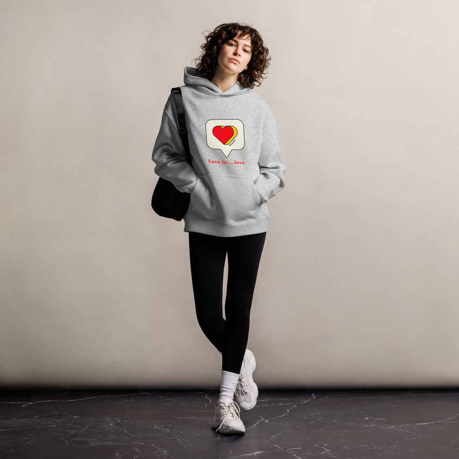 Woman wearing a gray 'Love is Love' hoodie with a heart graphic, standing confidently. Hoodie promotes inclusivity and love.